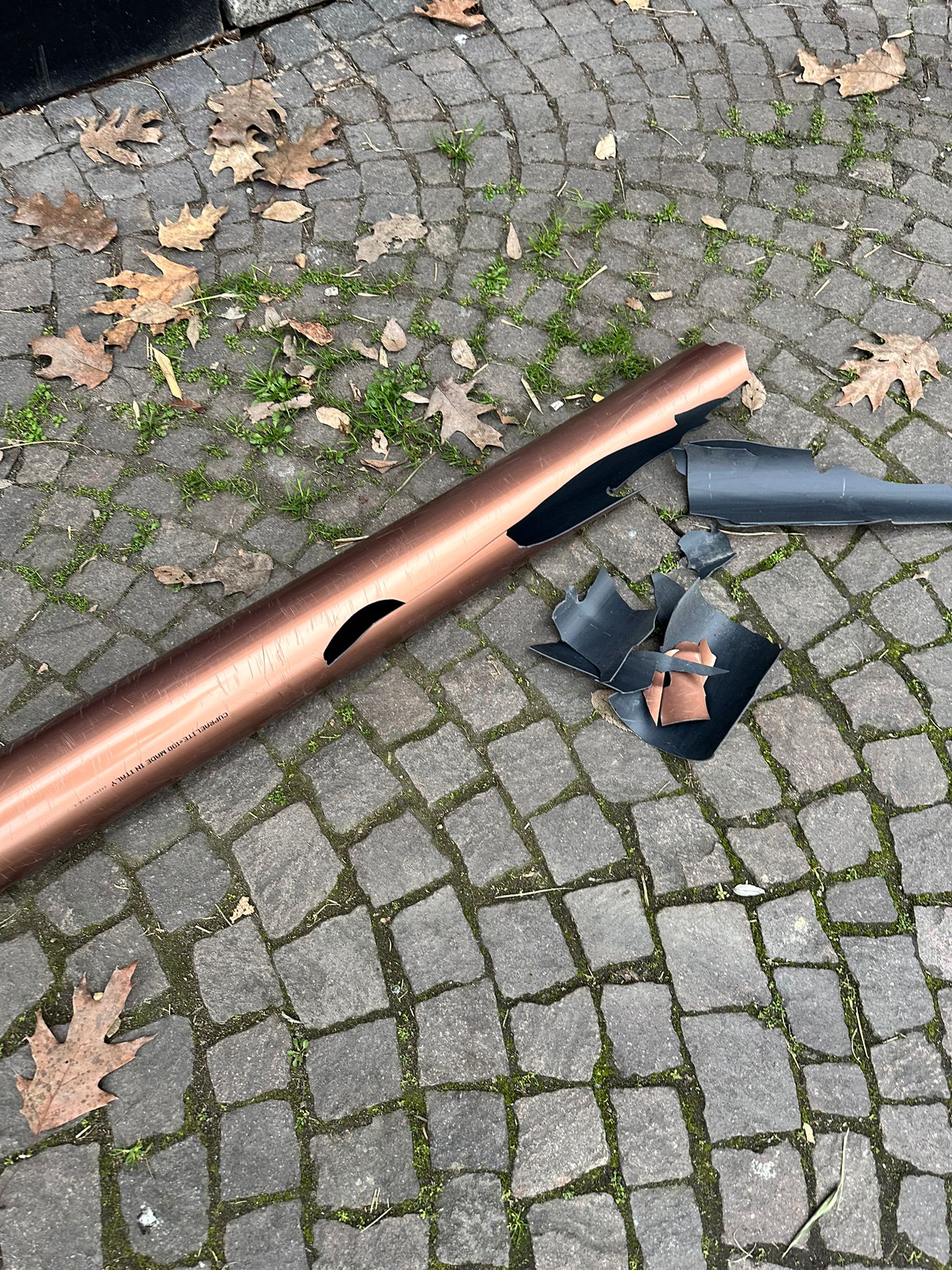 A broken copper-colored pipe and detached fittings lie on a mossy cobblestone surface.