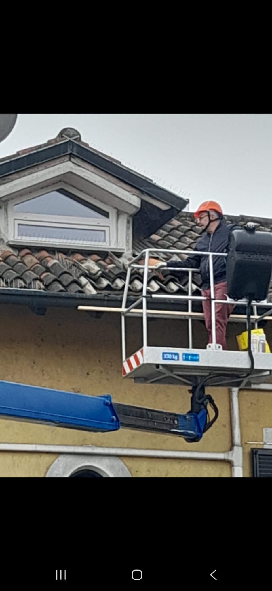 A worker in an orange helmet stands on an elevated platform beside a tiled roof under an overcast sky.