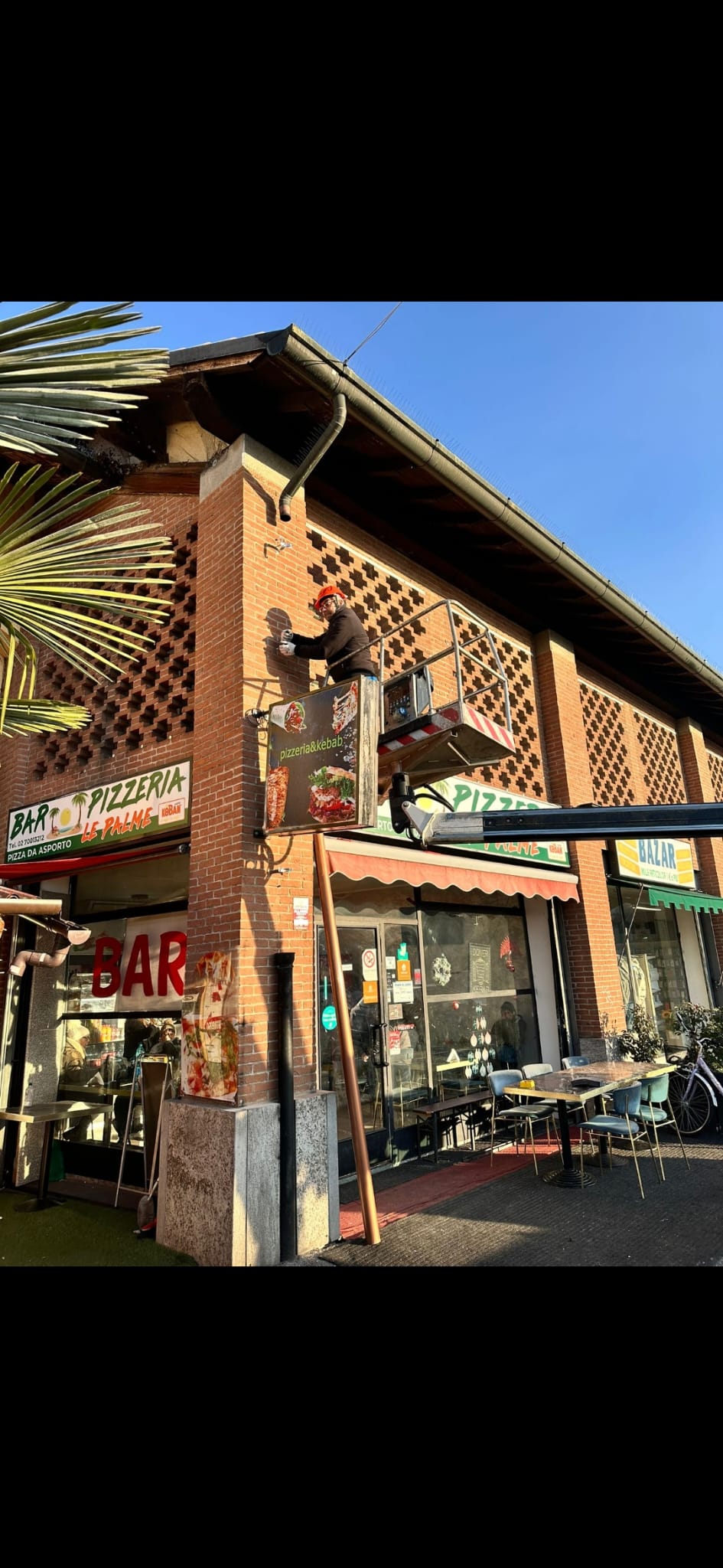 A worker on a raised platform works beside the upper facade of a brick-fronted building in bright sunlight.